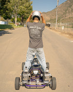 Person on a refurbished purple Steen's Pole-Cat Go-Kart vehicle with a helmet, standing on a road with trees and a mountain in the background.