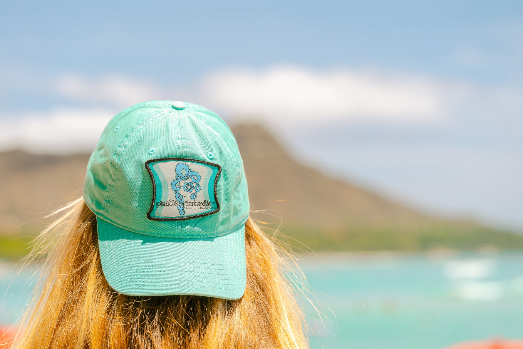 Surfer wearing a teal like color Plastic Fantastic hat with Diamond head in the background. 