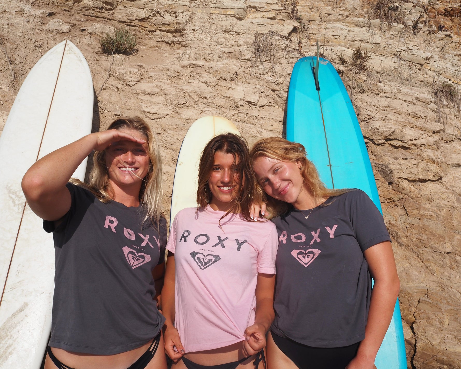 Three women with surfboards posing against a rocky wall, wearing Roxy shirts.